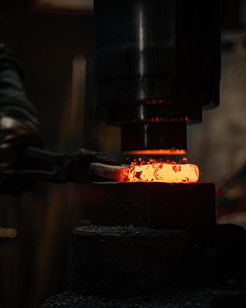 Blacksmith shaping red-hot iron with hammer under hydraulic press.